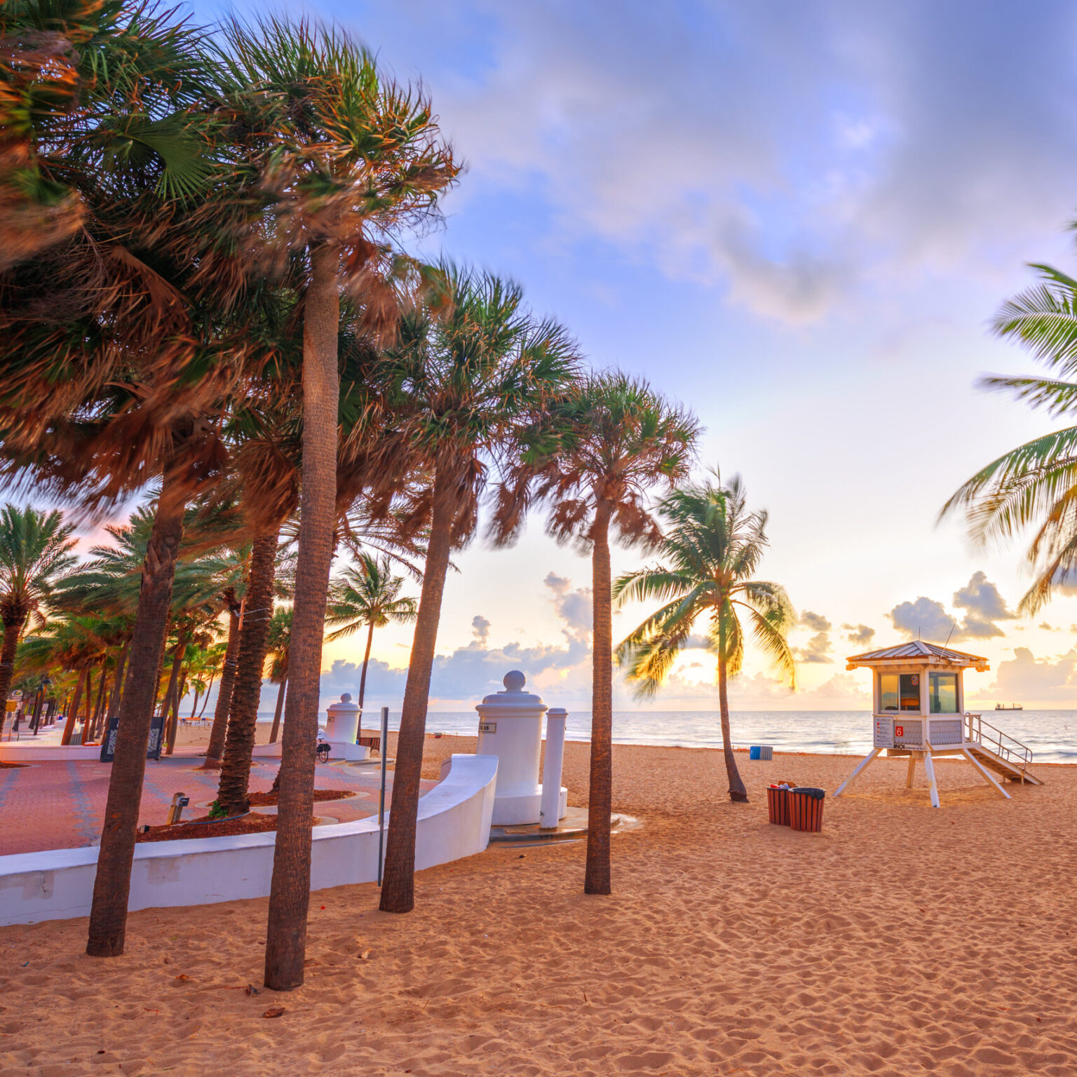 Sunrise over Fort Lauderdale Beach with palm trees, a lifeguard stand, and the signature white wave wall along the sandy shoreline in Florida.