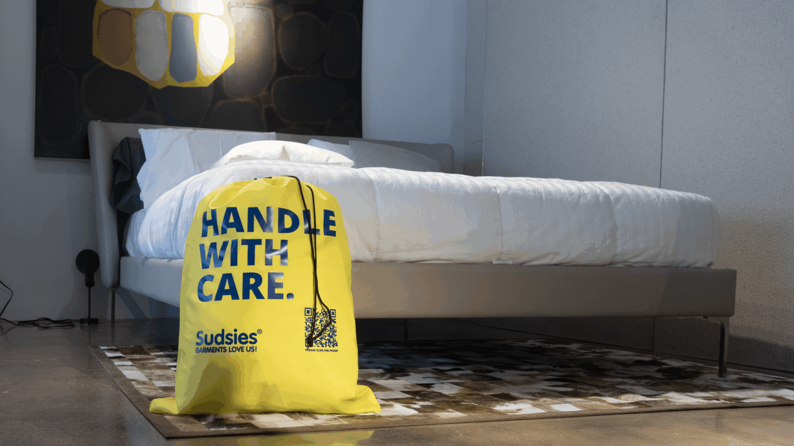 Bright yellow Sudsies laundry bag with “HANDLE WITH CARE” printed in bold blue letters, placed at the foot of a modern white bed in a stylish, minimal bedroom.