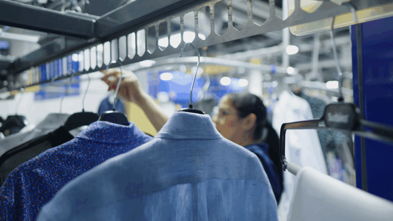Close-up of freshly cleaned garments on hangers moving along a conveyor system, with a Sudsies team member working in the background of a professional dry cleaning facility.
