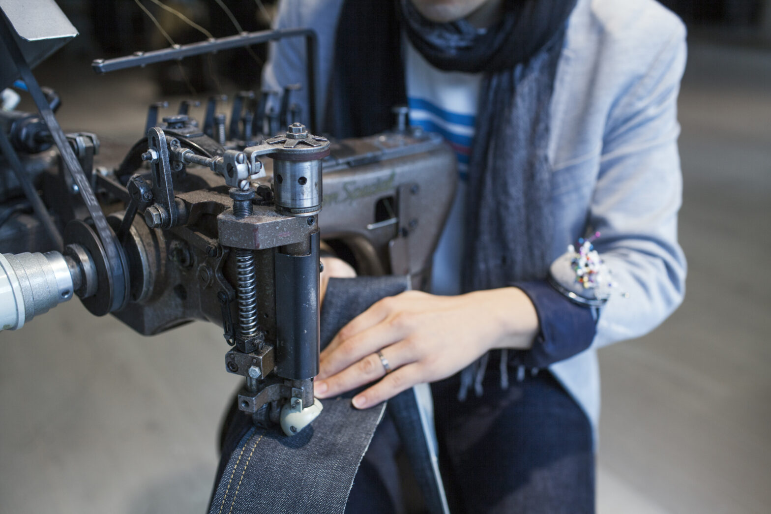 Close-up of a tailor using an industrial sewing machine to hem dark selvedge denim, wearing a wrist pin cushion and scarf, symbolizing expert garment craftsmanship.