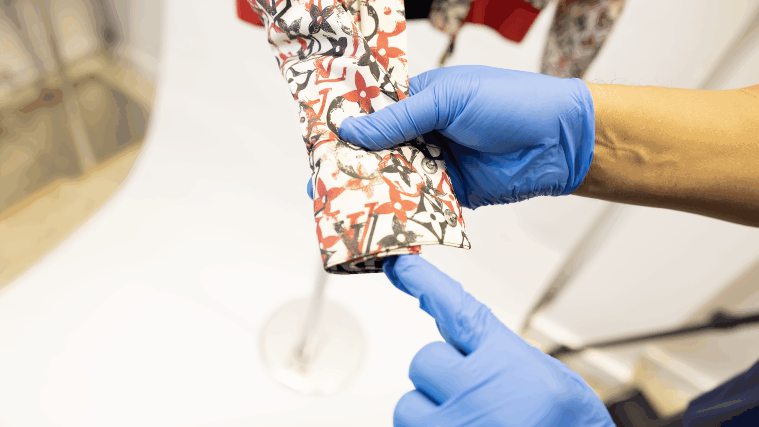 Close-up of a person wearing blue gloves inspecting the cuff of a designer Louis Vuitton shirt with red and black floral monogram print, demonstrating precision garment care.