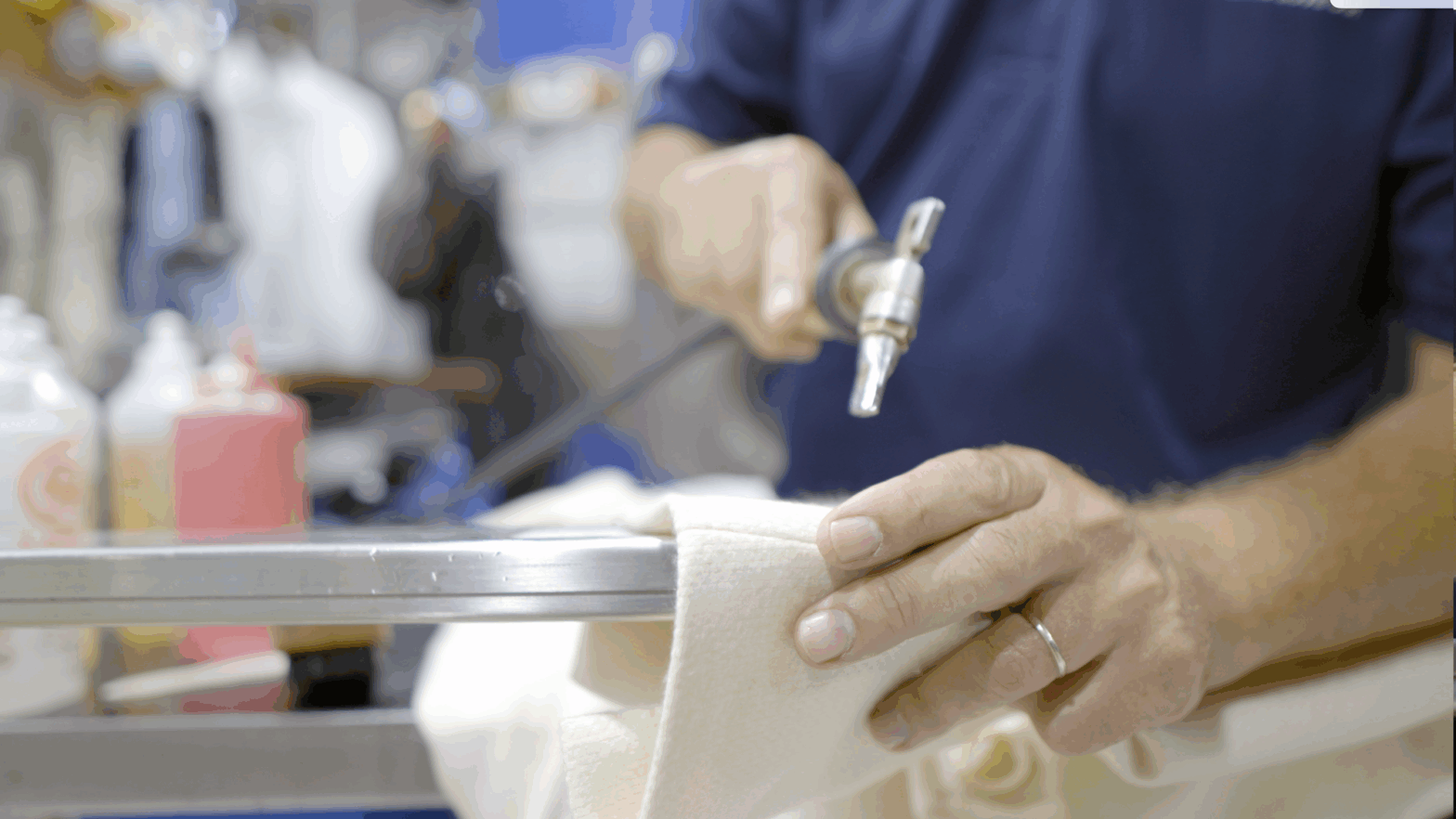 Close-up of a technician’s hands using a precision steam or air tool to spot-clean a delicate cream-colored garment, with cleaning agents and tools visible nearby.