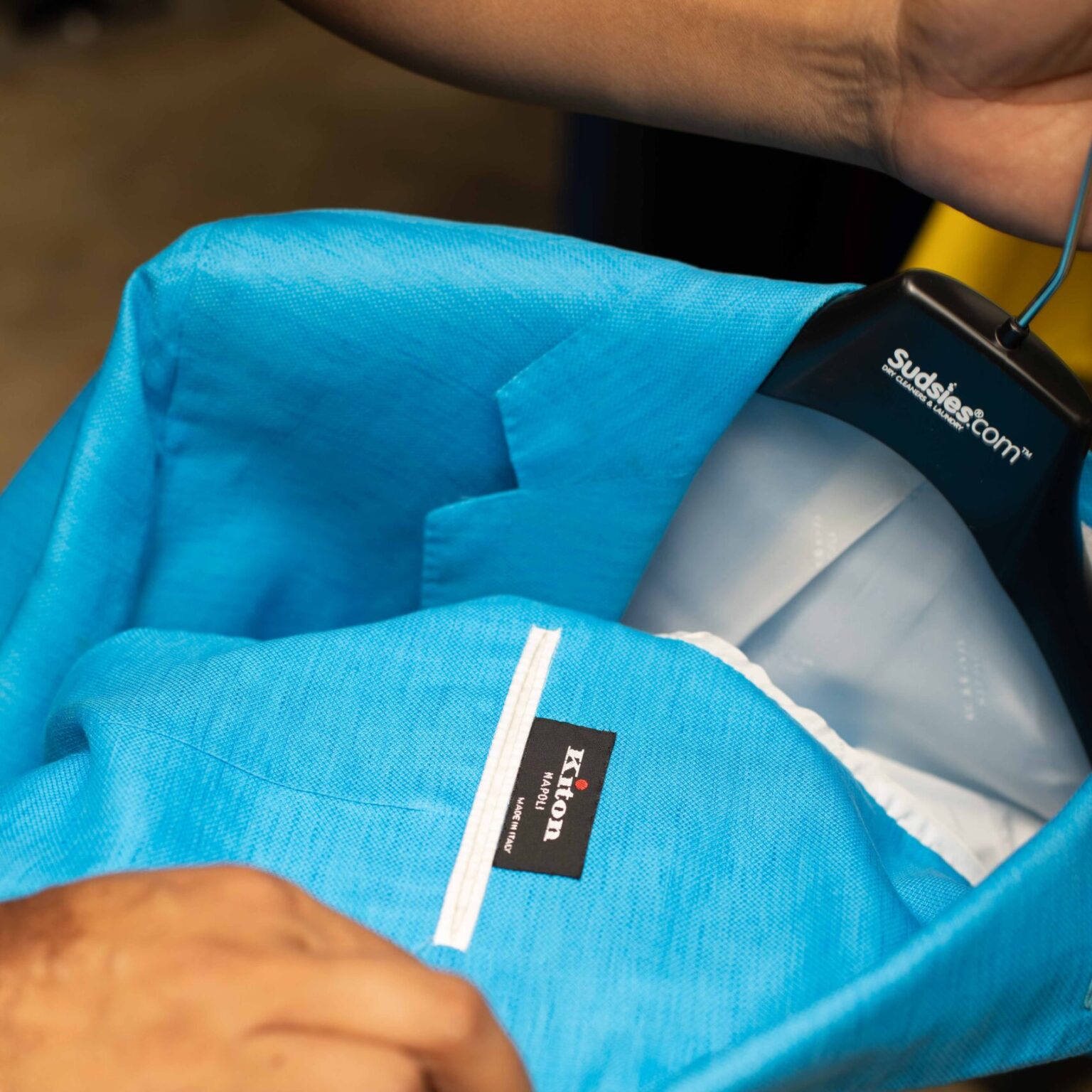 Close-up of a bright blue Kiton blazer on a black Sudsies.com™ hanger, showing the label and interior details being carefully handled by a technician.