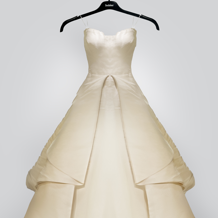 An elegant ivory ballgown-style wedding dress with layered satin and tulle, displayed on a black Sudsies hanger against a neutral gray background.