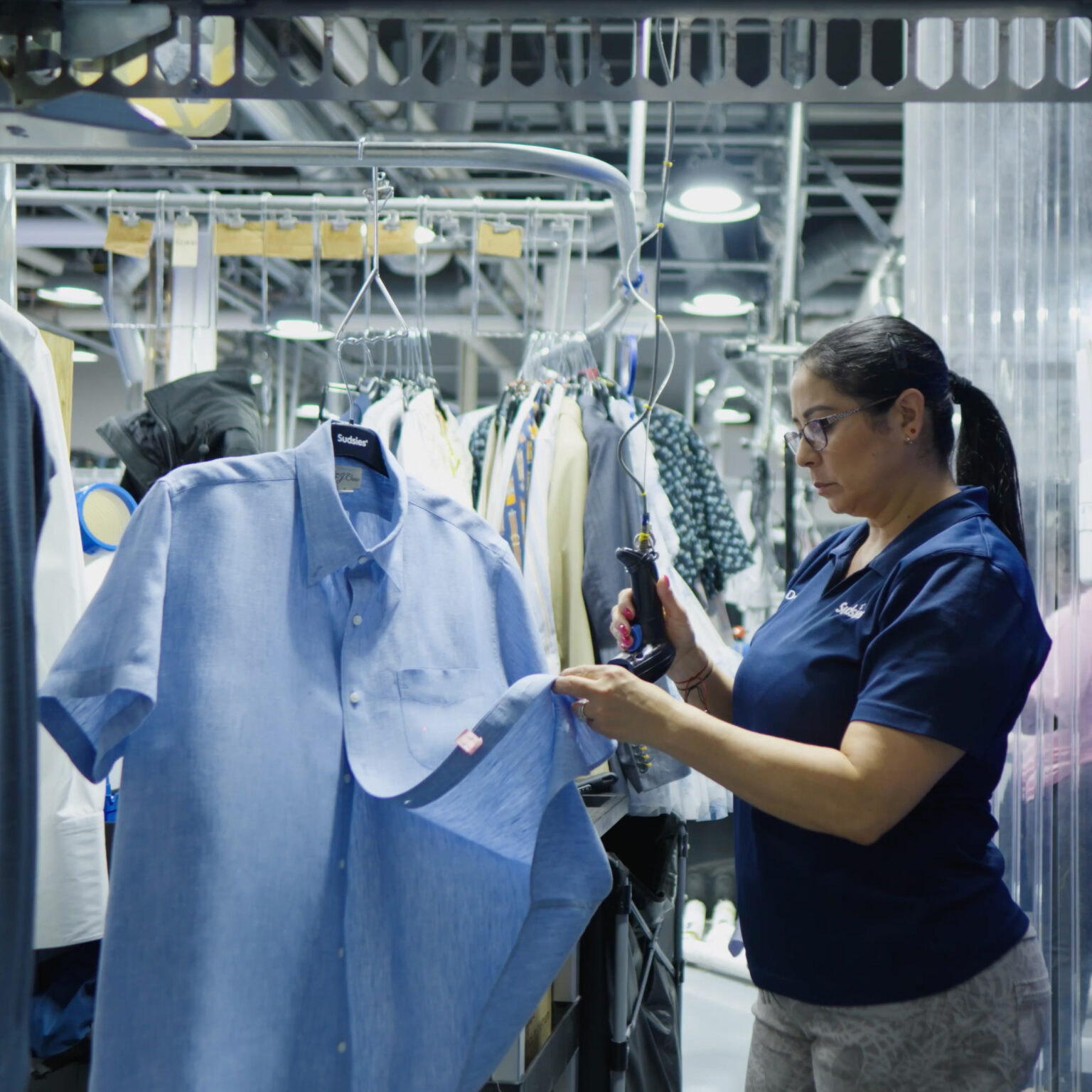 Sudsies technician in navy uniform inspects a freshly cleaned light blue short-sleeve shirt on a branded hanger beneath an overhead conveyor, surrounded by other garments.