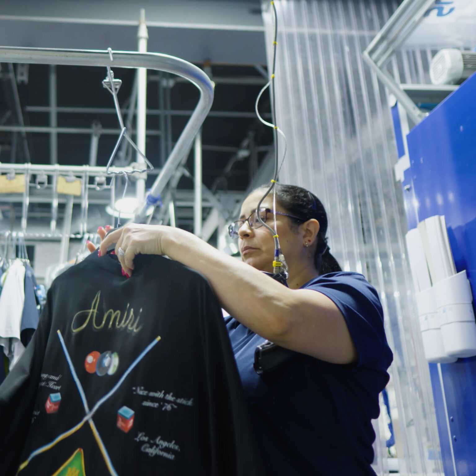 Sudsies technician carefully inspecting a designer Amiri shirt under garment conveyor lighting in a professional dry cleaning facility.