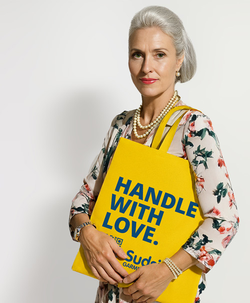 Elegant woman with silver hair holding a bright yellow Sudsies dry cleaning tote that reads “HANDLE WITH LOVE.”