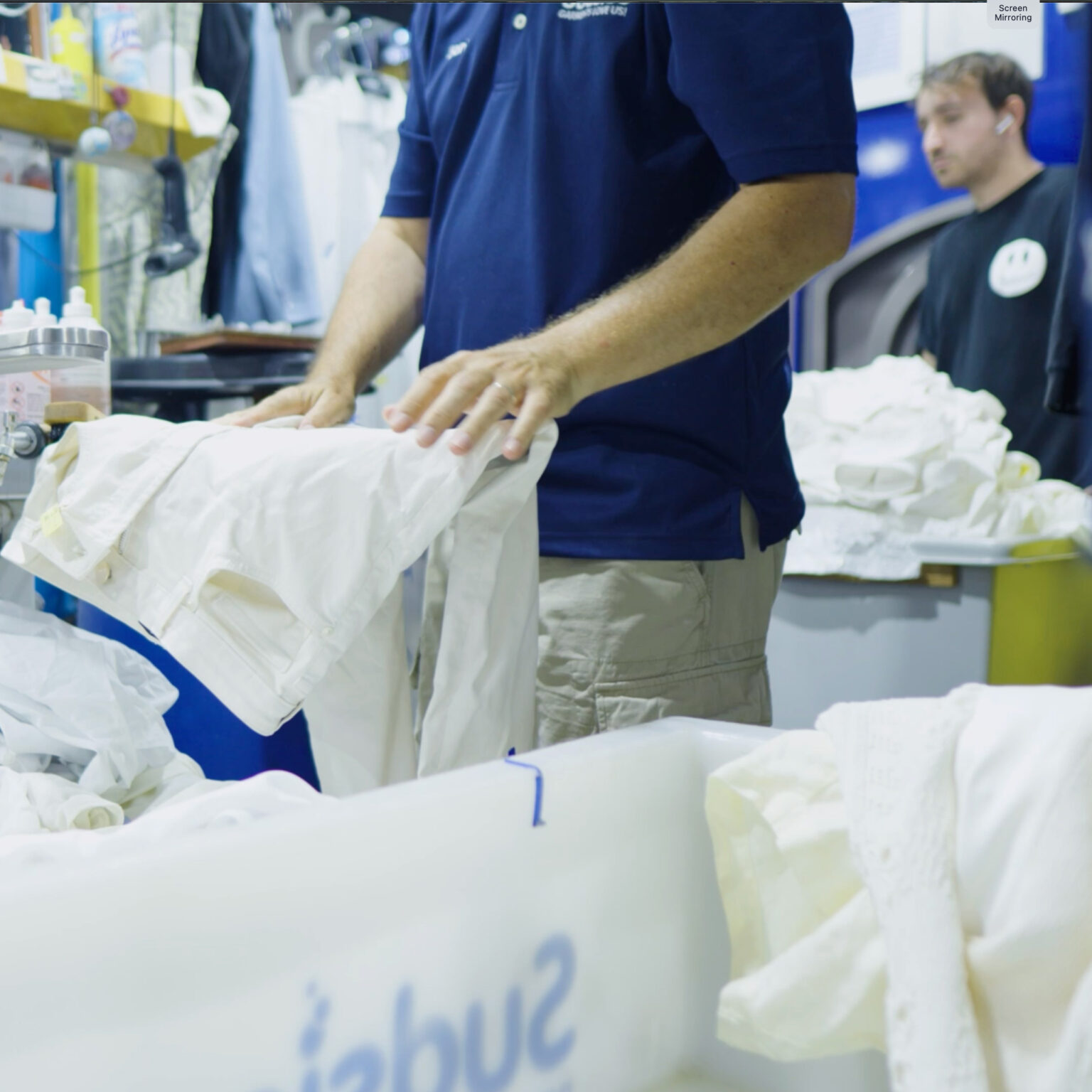 Technician in navy Sudsies polo shirt sorting or inspecting a white shirt in a garment processing area, surrounded by bins of clothing and cleaning supplies.
